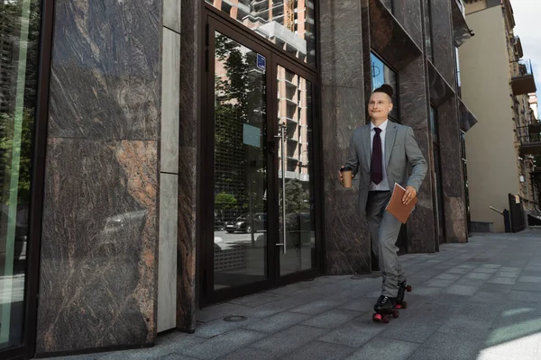 Smiling man in suit and roller skates riding with coffee to go along building on urban street — Stock Photo