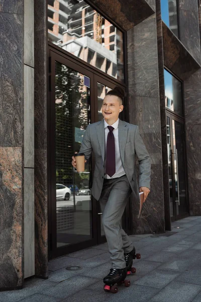 Счастливый человек в деловом костюме катается на роликах с бумажной чашкой и папкой на городской улице — стоковое фото