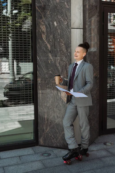 Homem de negócios sorridente em patins de patins de pé com documentos e café para ir perto de edifício na rua — Fotografia de Stock