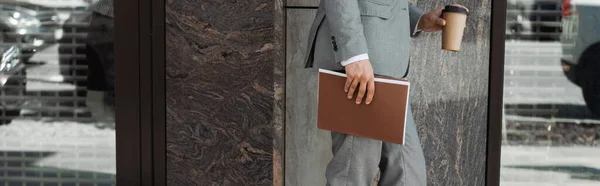 Vista parcial del hombre de negocios en traje de pie con bebida para llevar y carpeta cerca del edificio de la ciudad, pancarta - foto de stock