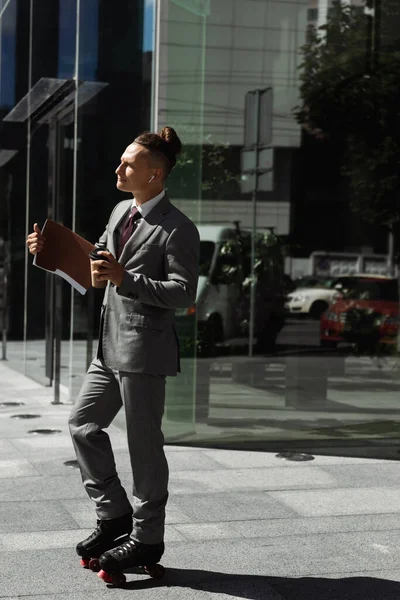 Стильный бизнесмен в роликовых коньках стоя с кофе идти и папки рядом с современным зданием в городе — стоковое фото