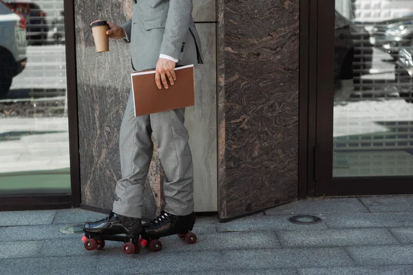 Vista recortada del patinador en traje de pie con bebida para llevar y carpeta cerca del edificio en la ciudad - foto de stock