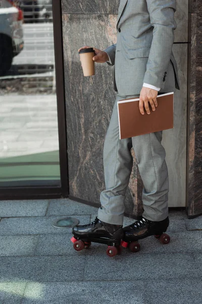 Vista cortada de empresário em patins de patins de pé com pasta e bebida takeaway perto de edifício na rua — Fotografia de Stock