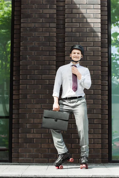 Cheerful roller skater in formal wear fixing tie and smiling at camera near building on street — Stock Photo