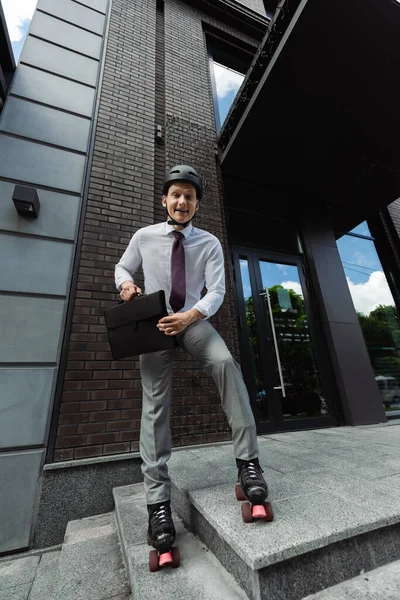Cheerful roller skater in formal wear and helmet holding briefcase and smiling at camera near modern building — Stock Photo