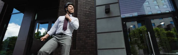 Низький кут зору бізнесмена в шоломі і формальний одяг, що стоїть з портфелем і фіксує краватку на вулиці, банер — стокове фото
