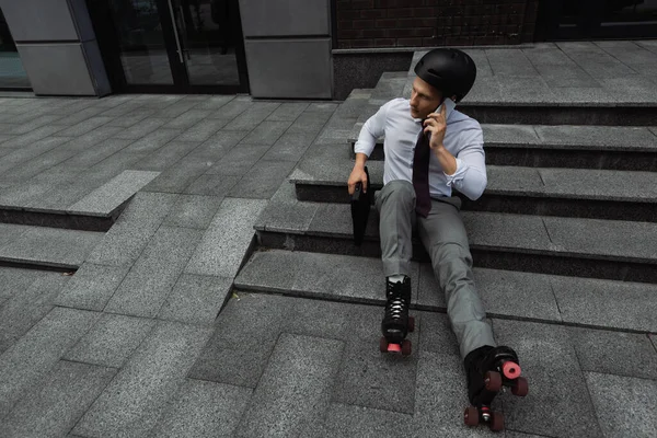 Blick aus der Vogelperspektive auf Geschäftsmann mit Helm und Rollschuhen, der auf dem Smartphone spricht, während er im Freien auf der Treppe sitzt — Stockfoto