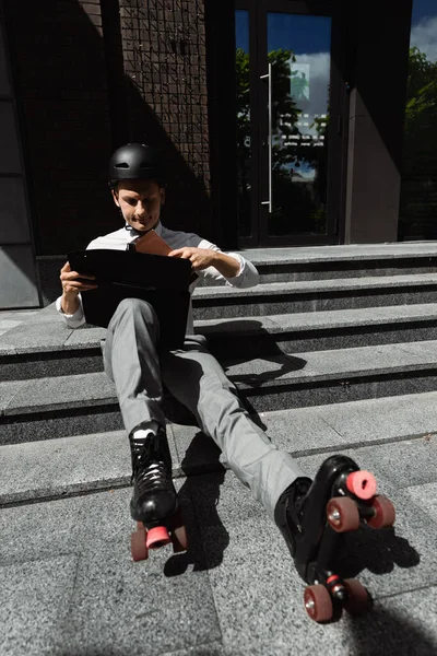 Улыбающийся бизнесмен в шлемах и роликовых коньках сидит на лестнице с портфелем и папкой — стоковое фото