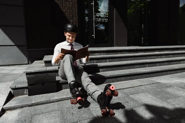Giovane uomo d'affari in pattini a rotelle e casco guardando il documento mentre seduto sulle scale all'aperto — Foto stock