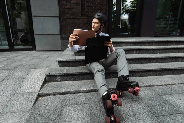 Бизнесмен в роликовых коньках смотрит в сторону, сидя на лестнице с портфелем и документами — стоковое фото