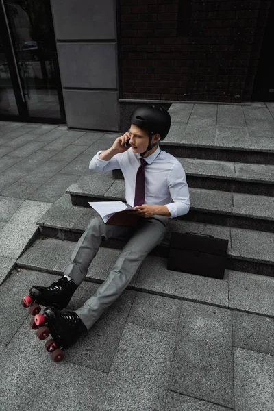 Roller skater in formal wear and helmet holding documents and talking on cellphone on stairs of city street — Stock Photo