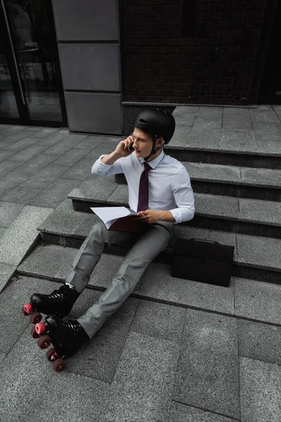 Patinador de ruedas en ropa formal y casco que sostiene documentos y habla por teléfono celular en las escaleras de la calle de la ciudad - foto de stock