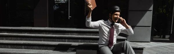Веселий бізнесмен в шоломі тримає папку в піднятій руці і розмовляє на мобільному телефоні на сходах на відкритому повітрі, банер — стокове фото