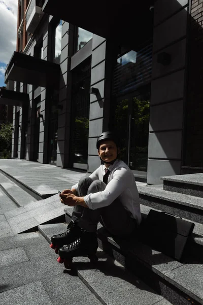 Homme d'affaires heureux en patins à roulettes et casque tenant smartphone et souriant à la caméra tout en étant assis sur les escaliers près du bâtiment — Photo de stock