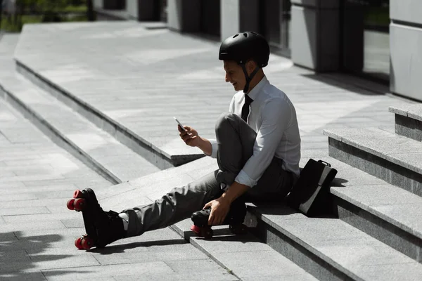 Усміхнений каток у формальному носінні та шоломі обміну повідомленнями на смартфоні, сидячи на міській вулиці — стокове фото