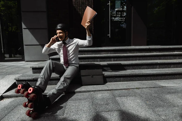 Hombre feliz en desgaste formal y patines de ruedas hablando en el teléfono inteligente mientras está sentado en las escaleras con la carpeta en la mano levantada - foto de stock
