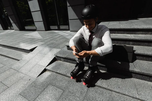 Vista de ángulo alto del hombre de negocios en patines de ruedas sentado en las escaleras y mensajería en el teléfono inteligente - foto de stock