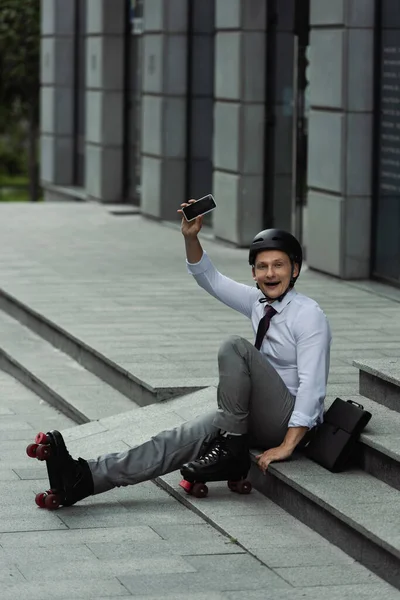 Aufgeregter Geschäftsmann mit Helm und Rollschuhen, Smartphone mit leerem Bildschirm in der Hand, während er draußen auf der Treppe sitzt — Stockfoto
