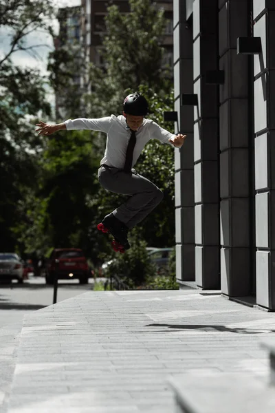 Comprimento total do homem no desgaste formal e patins com capacete pulando ao fazer truque na rua urbana — Fotografia de Stock