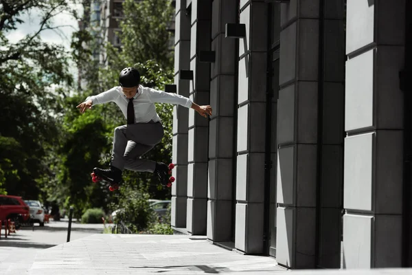 Longitud completa de hombre de negocios en casco y patines saltando mientras hace truco en la calle de la ciudad - foto de stock