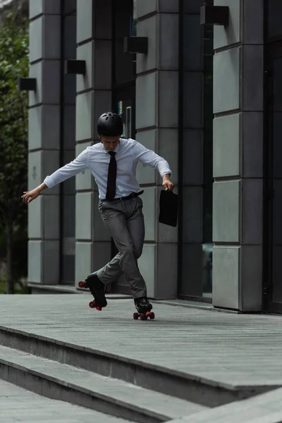 Мужчина в формальном костюме и на роликовых коньках выполняет трюк возле современного здания на городской улице — стоковое фото