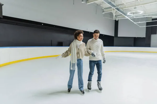 Joven pareja interracial cogida de la mano mientras patina sobre hielo en pista de patinaje - foto de stock