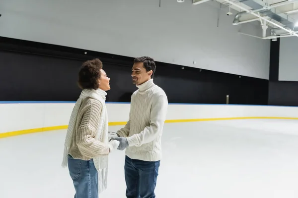 Multiethnic couple in warm knitted sweaters holding hands on ice rink — Stock Photo