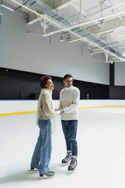 Positiva pareja multiétnica cogida de la mano y mirando hacia otro lado en pista de hielo - foto de stock
