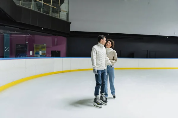 Pareja multiétnica positiva en suéteres calientes patinaje sobre hielo en pista de patinaje - foto de stock