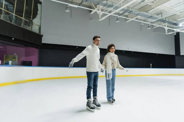 Feliz pareja interracial cogida de la mano mientras patina sobre hielo en pista de patinaje - foto de stock