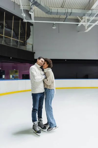 Багатоетнічна пара в теплих светрах і рукавичках обіймається на ковзанах — стокове фото