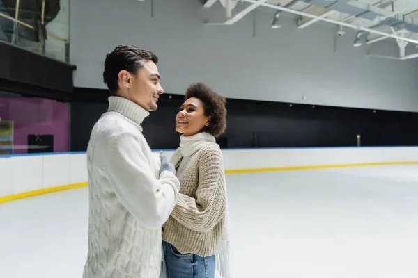 Sorridente donna africana americana guardando il fidanzato in maglione sulla pista di pattinaggio sul ghiaccio — Foto stock
