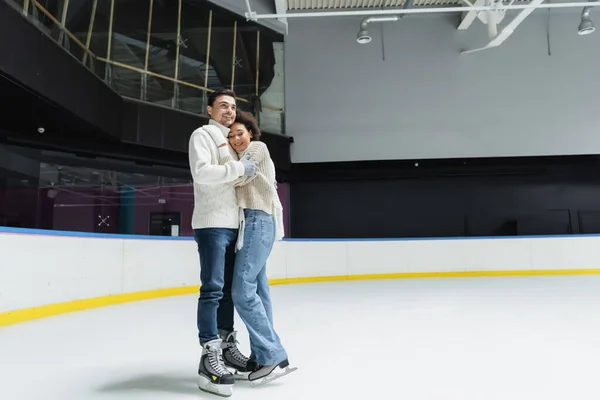 Усміхнений чоловік в светрі і ковзанах обіймає афро-американську дівчину на ковзанці — стокове фото