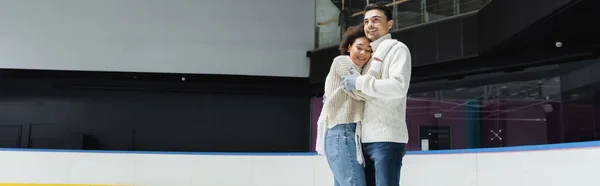 Hombre en jersey de punto abrazando a novia afroamericana en pista de hielo, pancarta - foto de stock