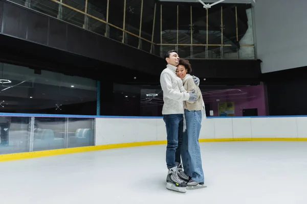 Hombre alegre abrazando a novia afroamericana en patines de hielo en pista de patinaje - foto de stock
