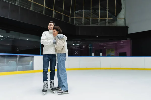 Smiling man hugging african american girlfriend in sweater and scarf on ice rink — Stock Photo