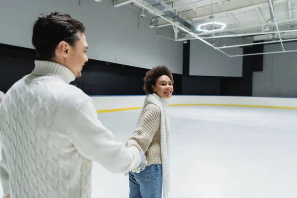 Smiling african american woman looking at camera while holding hand of boyfriend on ice rink — Stock Photo