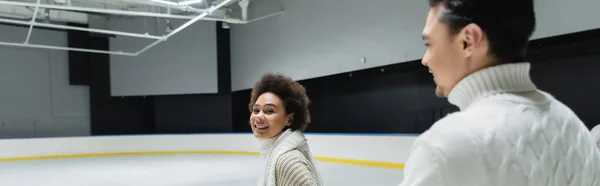 Young african american woman looking at camera near blurred boyfriend on ice rink, banner — Stock Photo