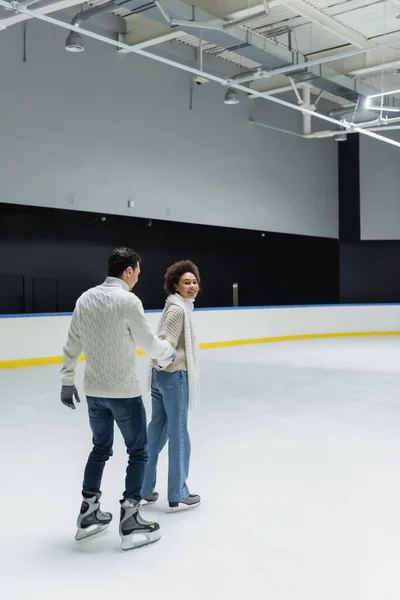 Alegre mujer afroamericana cogida de la mano del novio mientras patina sobre hielo en la pista - foto de stock