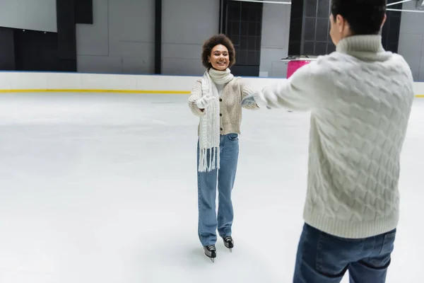 Cheerful african american woman outstretching hands to blurred boyfriend on ice rink — Stock Photo