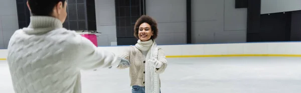 Mujer afroamericana joven extendiendo las manos a su novio en pista de hielo, pancarta - foto de stock