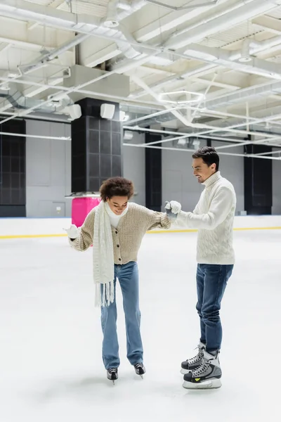 Smiling man helping to african american girlfriend skating on ice rink — Stock Photo