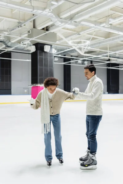 Un homme souriant aide une petite amie afro-américaine à patiner sur une patinoire — Photo de stock