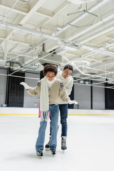Hombre positivo abrazando a novia afroamericana mientras patina sobre hielo en pista — Stock Photo