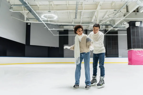 Cheerful man hugging and teaching african american girlfriend on ice rink — Stock Photo