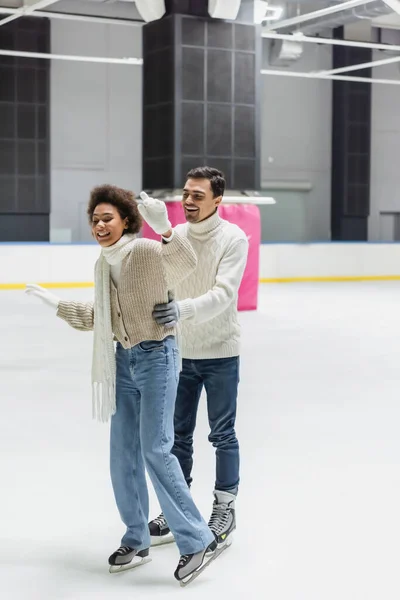 Young man in sweater hugging african american girlfriend skating on ice rink — Stock Photo