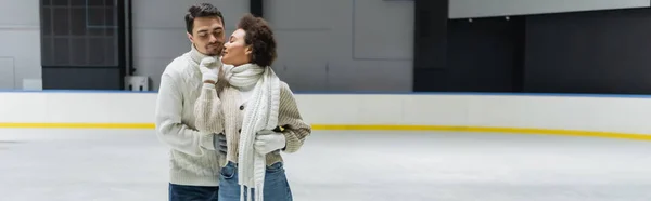 Mujer afroamericana en ropa de abrigo besando novio en suéter en pista de hielo, pancarta - foto de stock