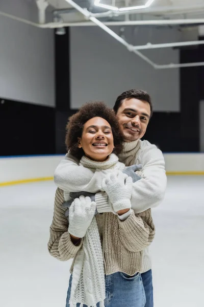 Hombre alegre abrazando a la novia afroamericana y mirando la cámara en la pista de hielo - foto de stock