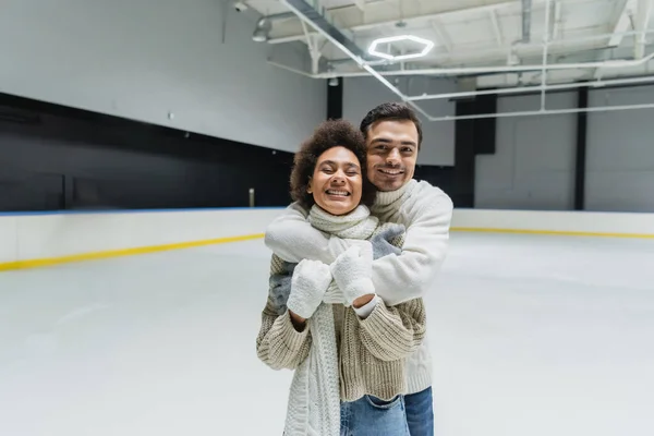 Hombre alegre en suéter y guantes abrazando a novia afroamericana mientras pasa tiempo en pista de hielo - foto de stock
