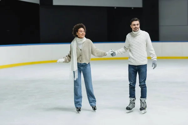Smiling multiethnic couple in sweaters and jeans skating during date on ice rink — Stock Photo
